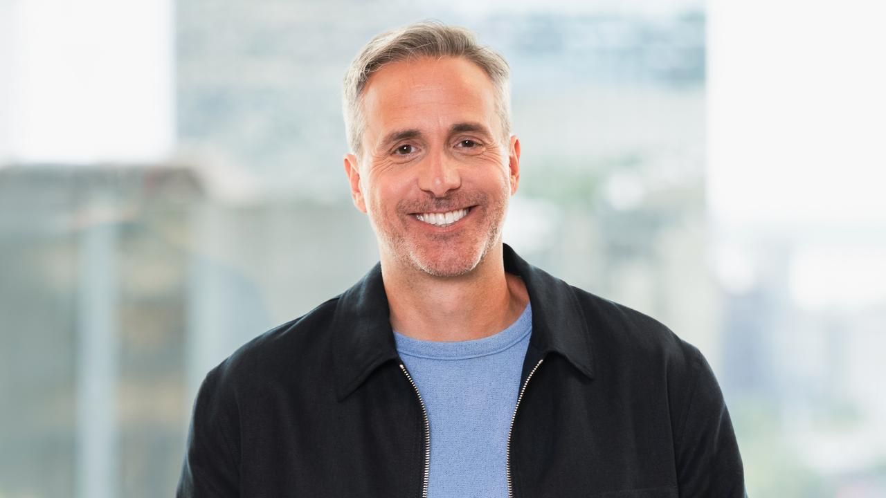 Dan Ayoub smiling in a black jacket and blue shirt in front of large window.